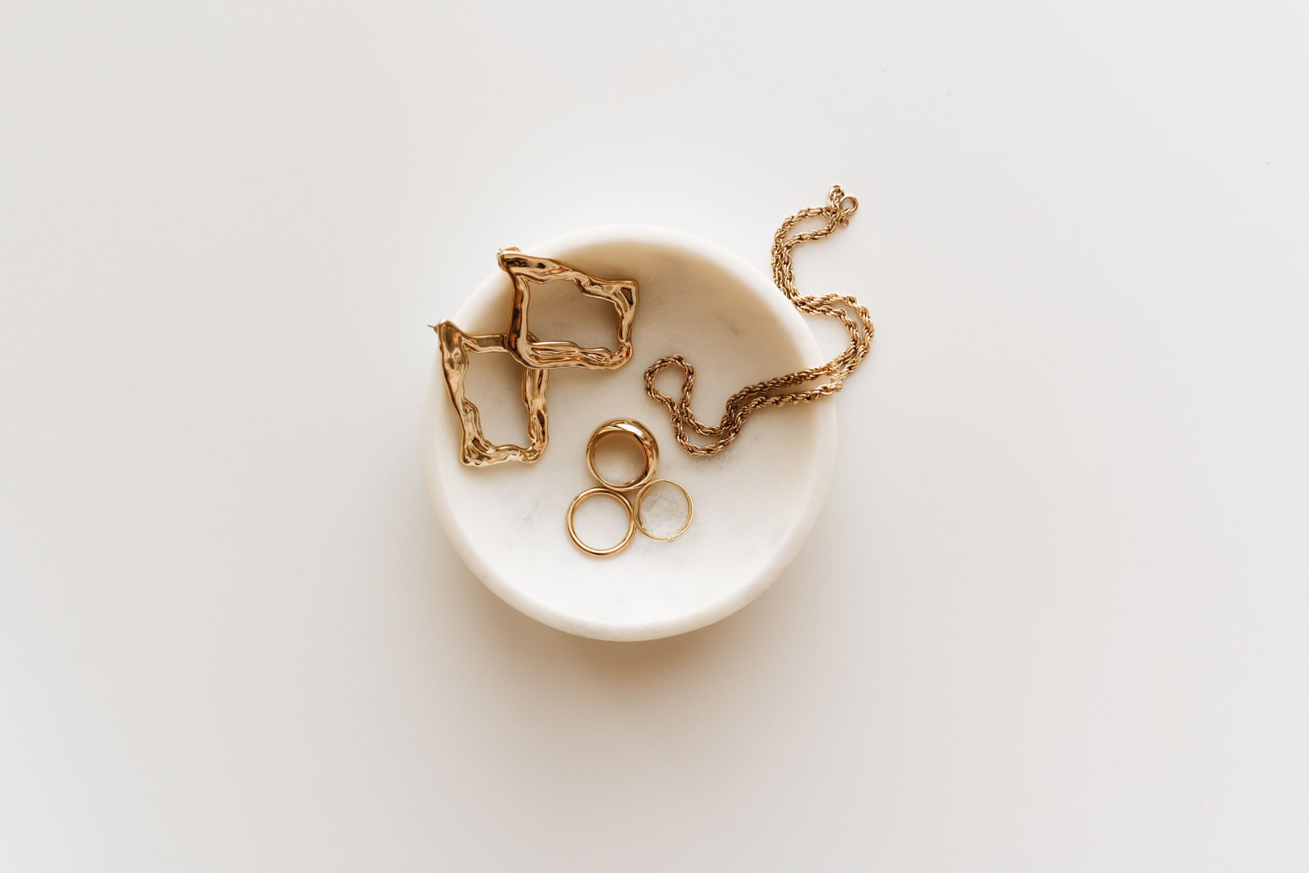 Stylish arrangement of gold jewelry including rings, earrings, and a necklace on a marble dish.