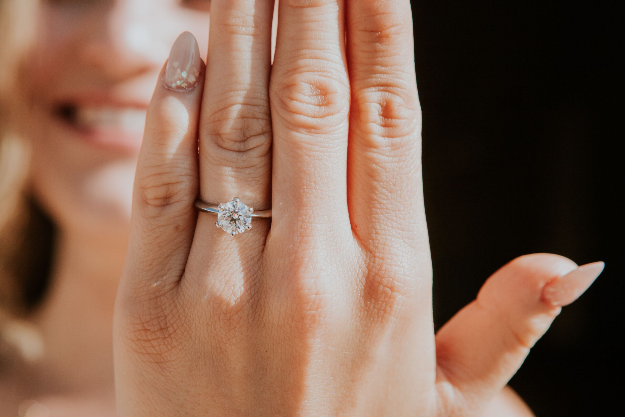 A close-up of a woman's hand displaying an elegant engagement ring with a smiling face in the background.