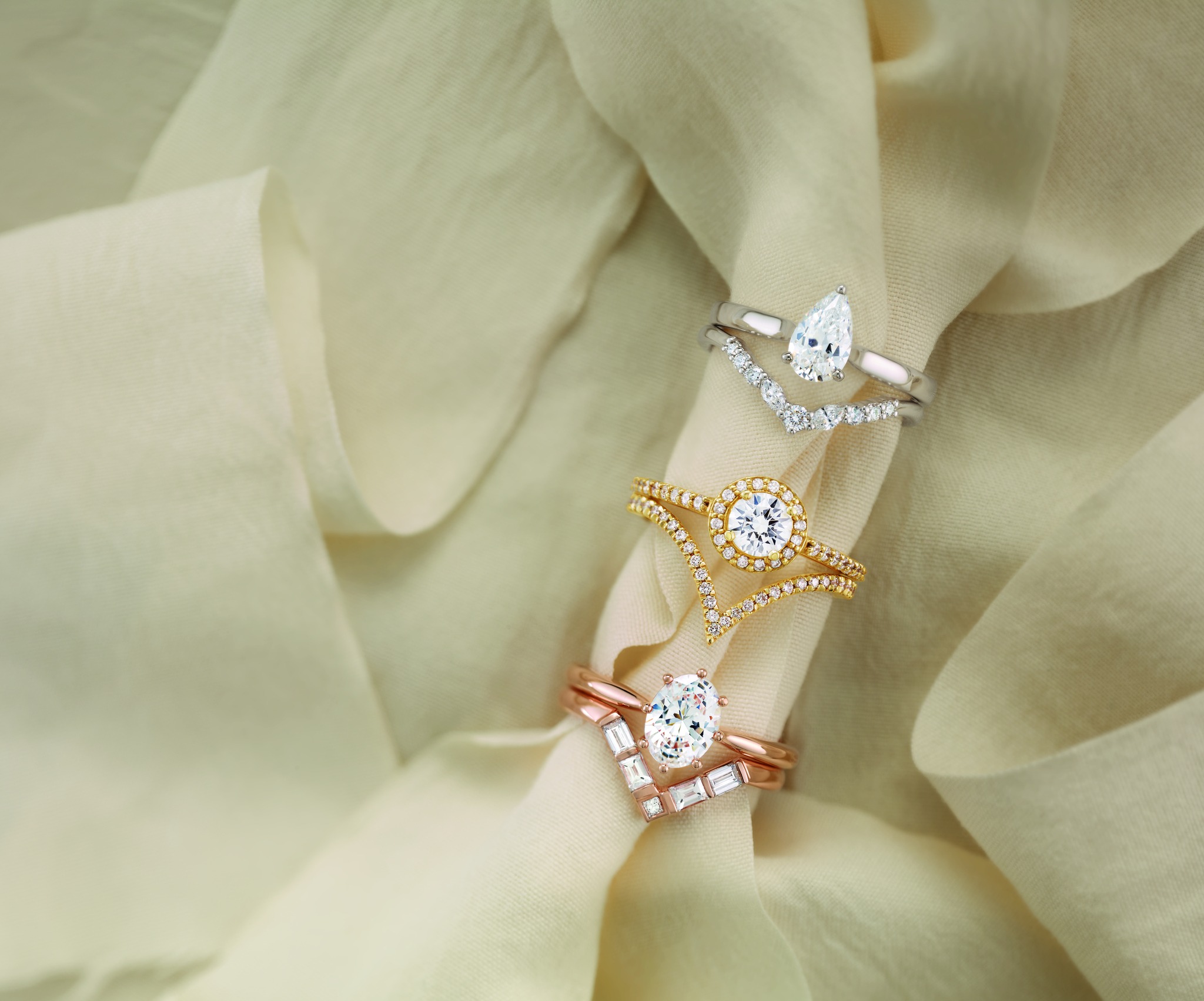 Three elegant rings set on a soft, beige fabric backdrop: a silver ring with a teardrop gem, a gold ring with a round gem, and a rose gold ring with a mixed-cut gem.